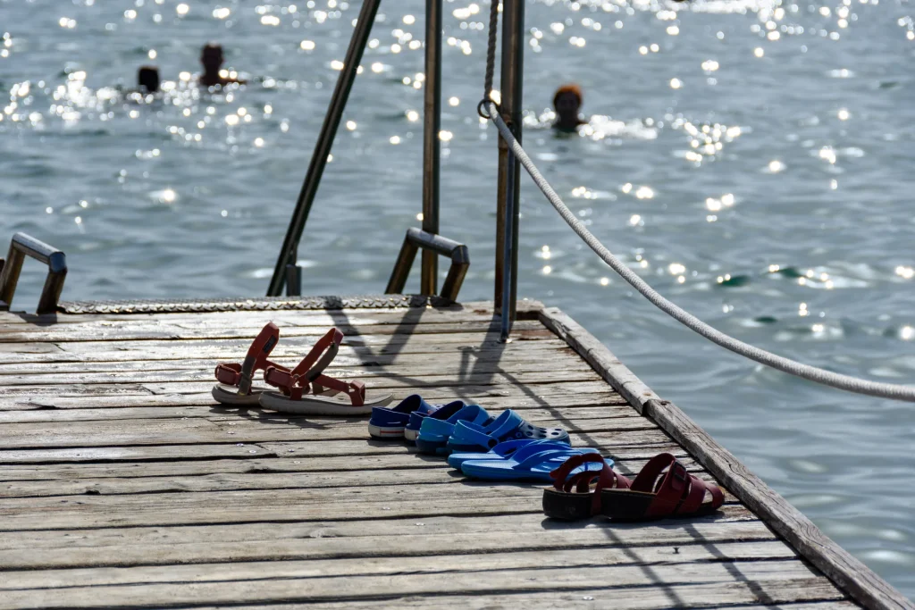 Sandales posées sur une jetée en bois au bord du lac d’Enghien-les-Bains, avec des baigneurs au loin, illustrant l’ambiance estivale des activités nautiques.
